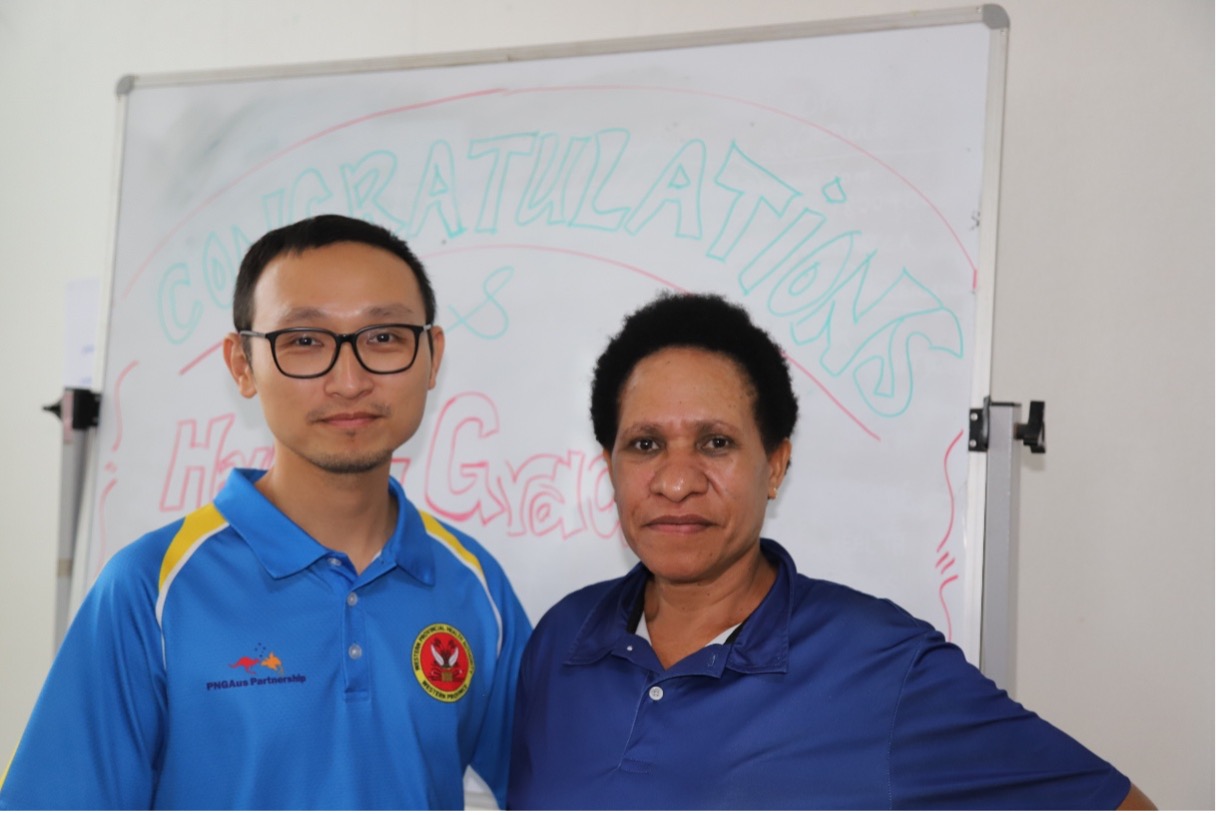 A man wearing glasses (left), and a woman with short hair (right), wearing blue polo shirts and standing in front of a white board.