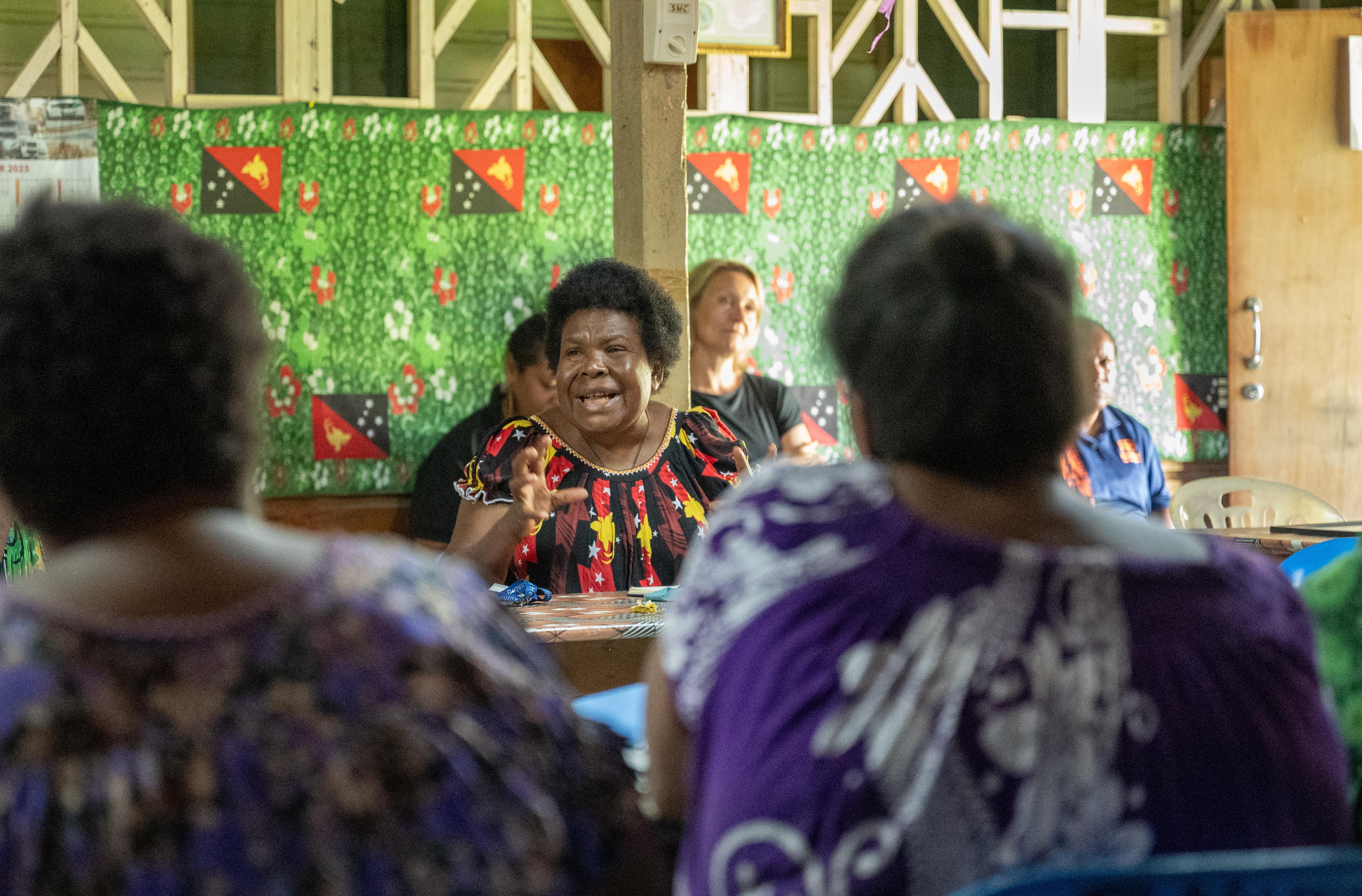 A woman speaks animatedly to a small group seated around a table inside a community hall decorated with Papua New Guinea flags, while others listen attentively in the foreground.