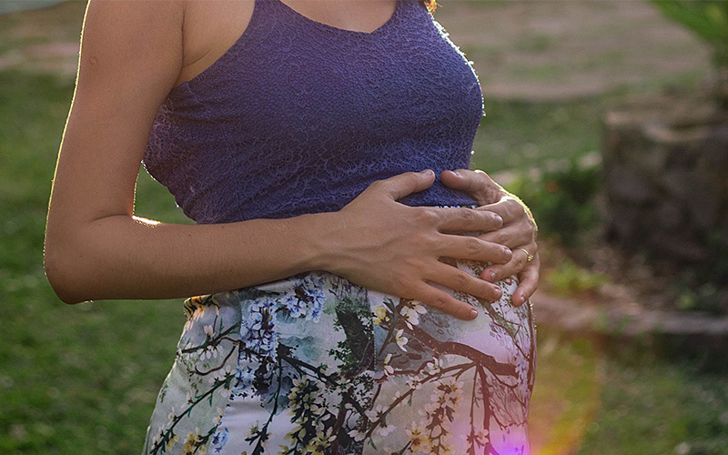A woman wearing a purple top and a floral skirt holding her pregnant belly.
