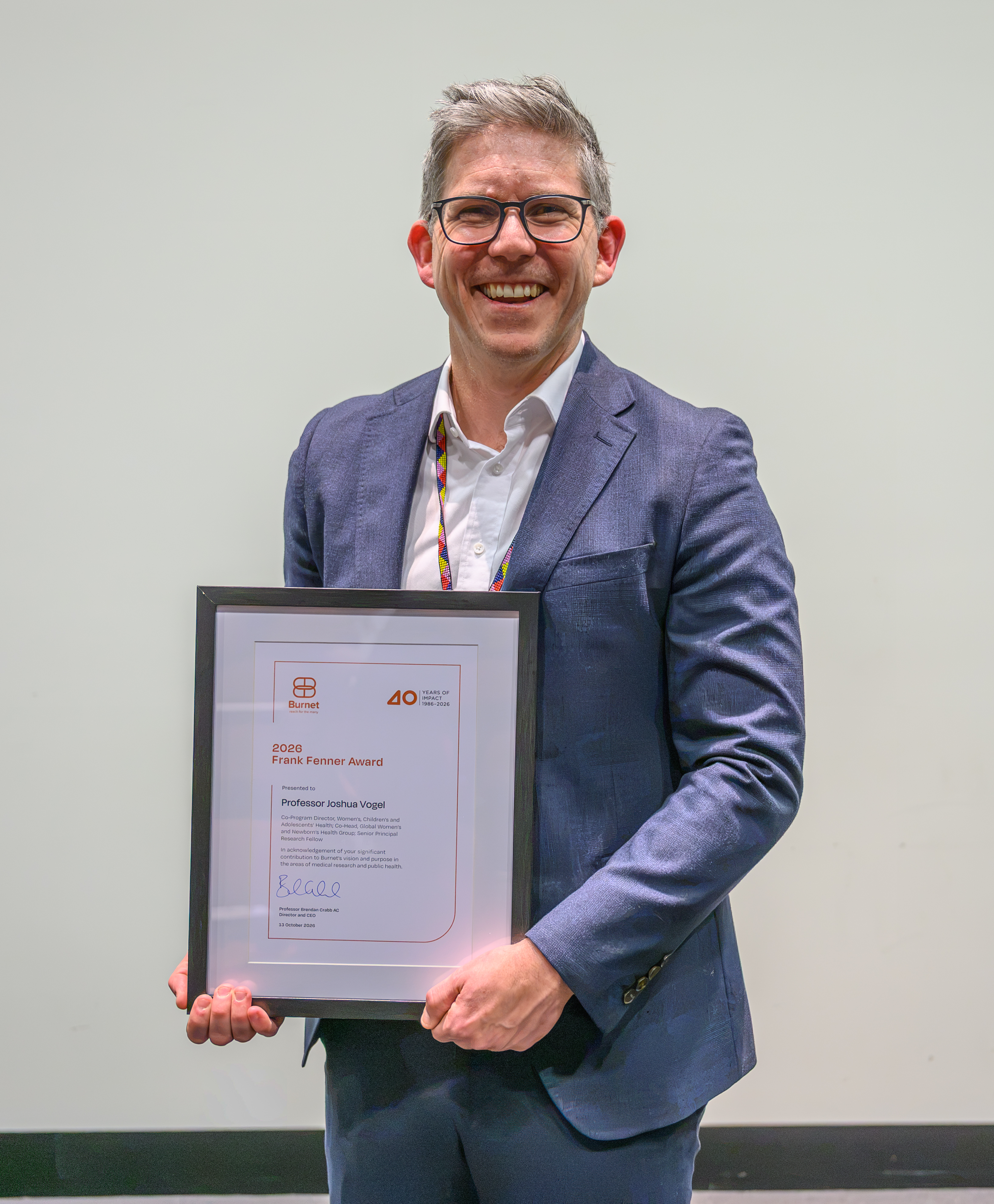 Professor Joshua Vogel smiles while holding his framed 2026 Frank Fenner Award at Burnet Institute, wearing a blue suit jacket and white shirt.