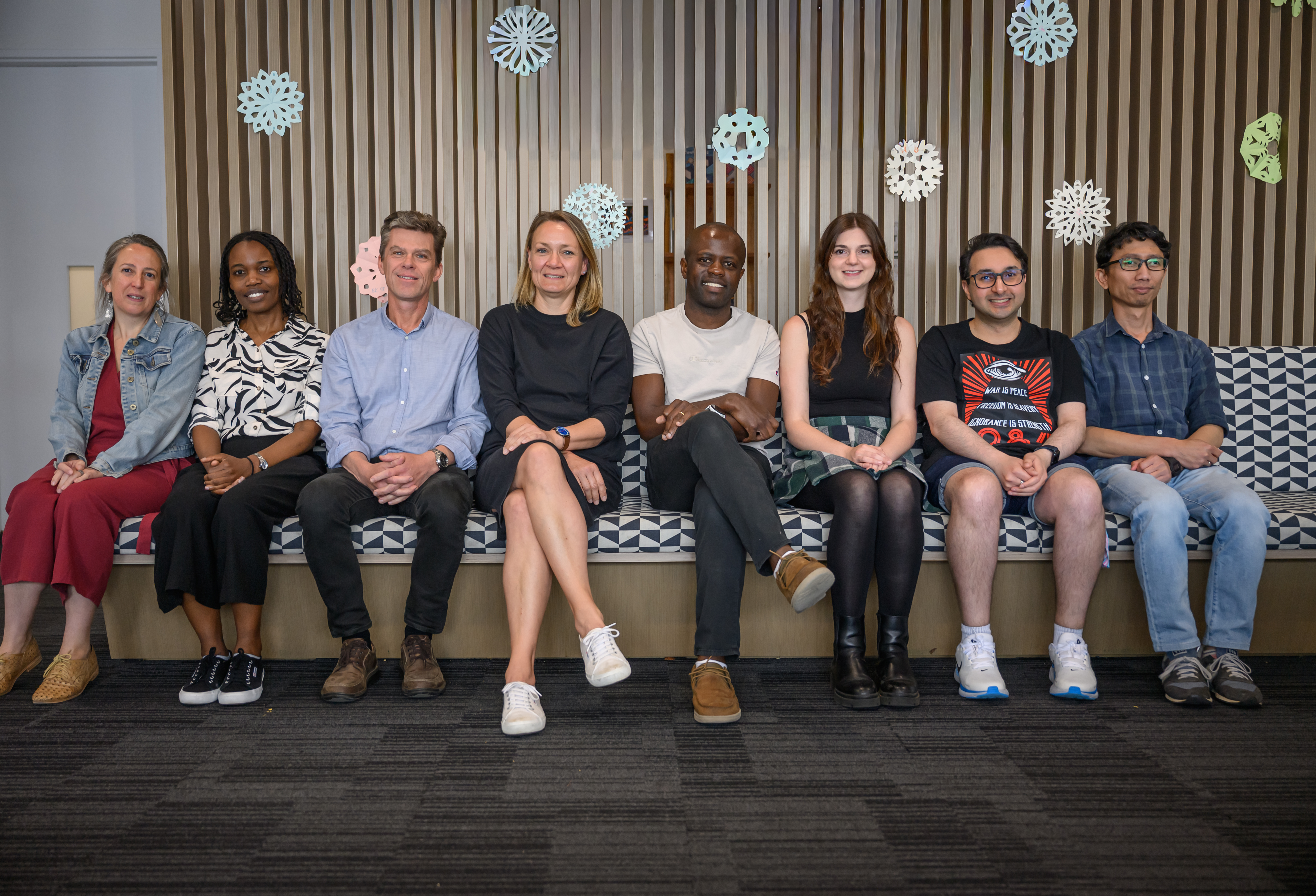 Eight researchers seated side by side on a patterned bench in a modern indoor space, smiling toward the camera. They are dressed in smart-casual clothing, with decorative paper snowflakes displayed on a wooden slat wall behind them.