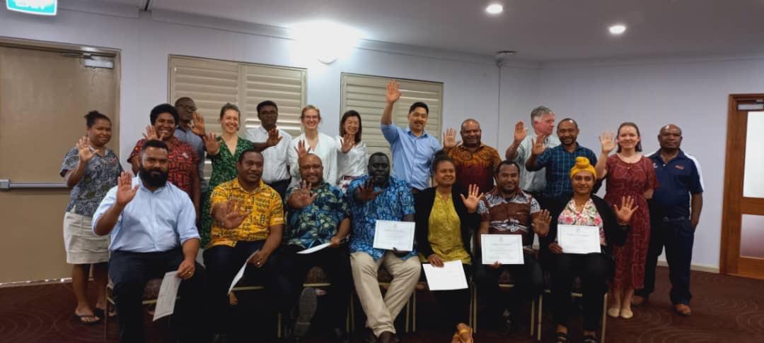 Two rows of people in a room. Front row of people with certificates and back row standing up. They have their palms open towards the camera.