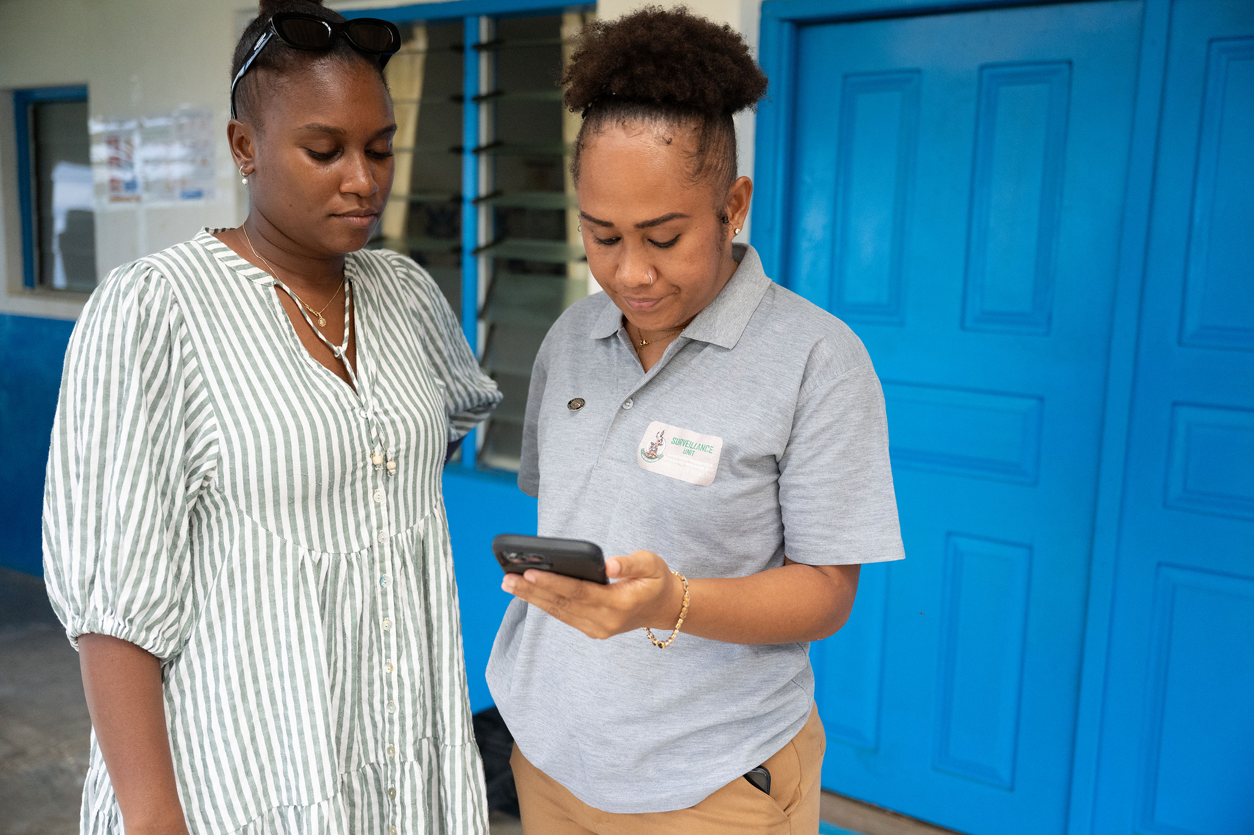 Two healthcare workers stand outside in front of a blue door. One holds a mobile phone. Both are looking at the phone. 