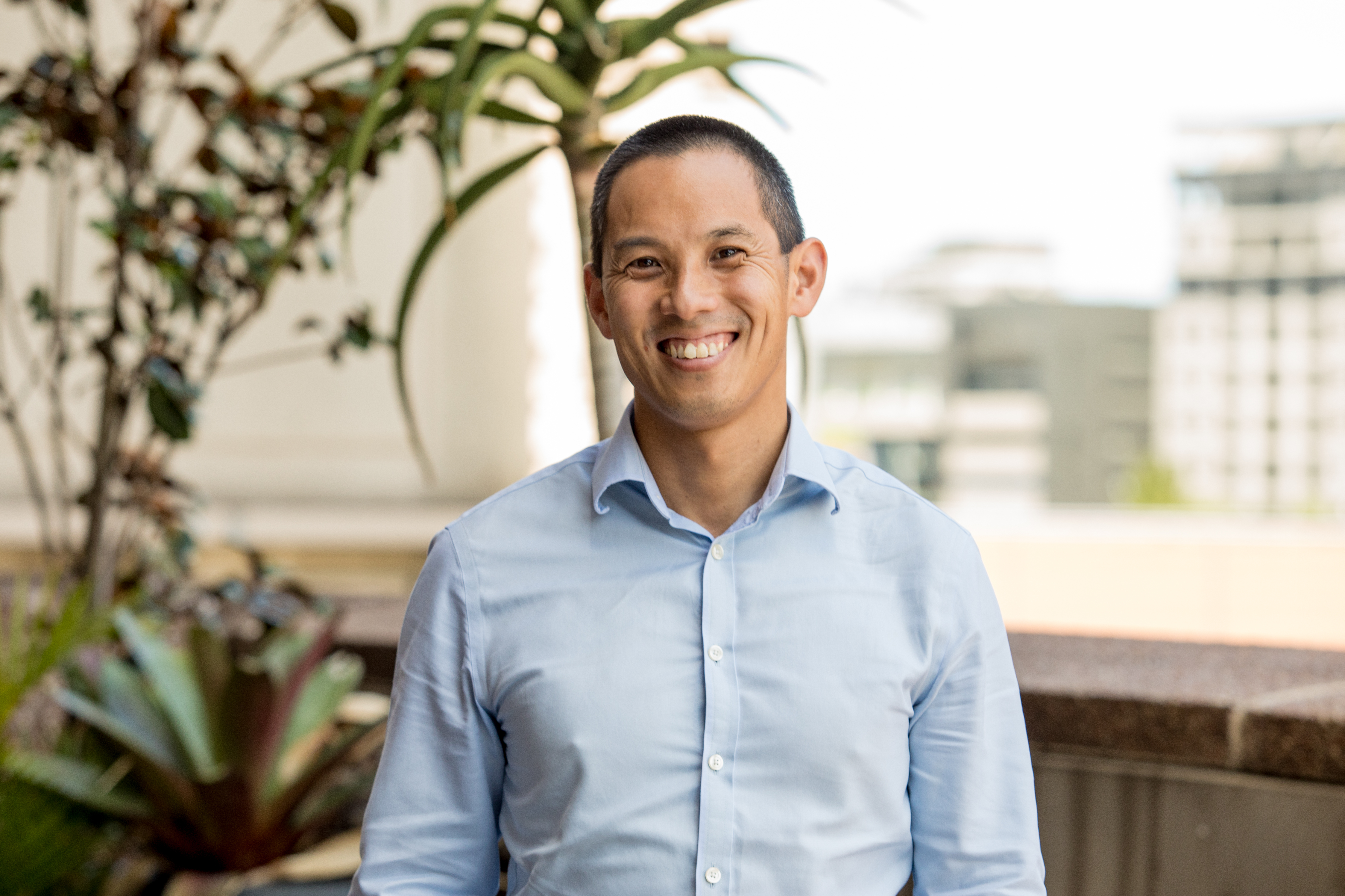 Portrait of Roger Chao wearing a blue shirt. He's standing on a balcony with a cityscape in the background.