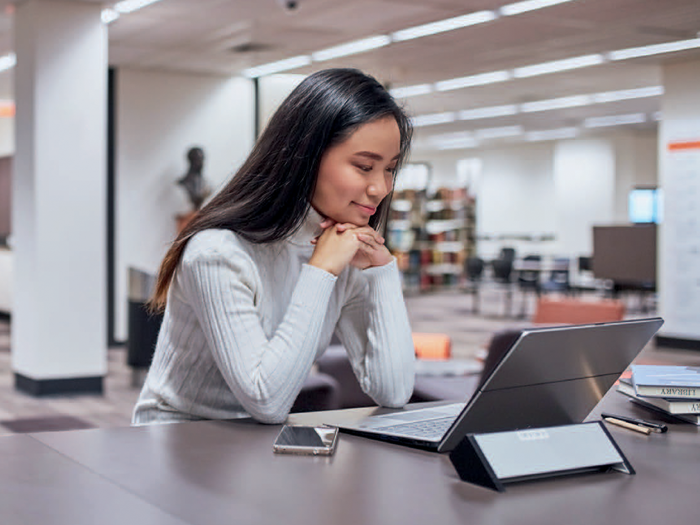 A student smiling at a laptop in a library.