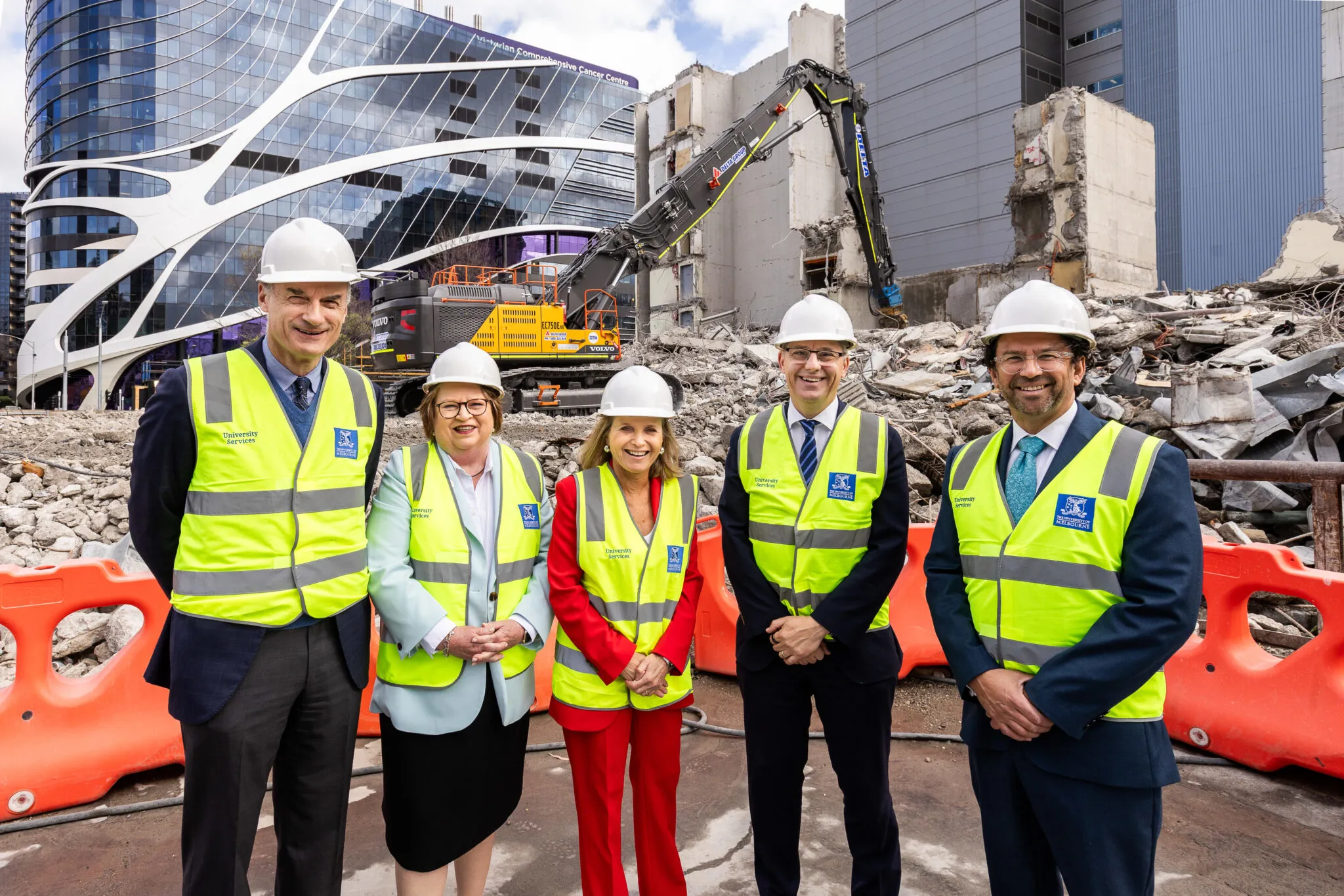 Five people wearing white hard hats and bright yellow University of Melbourne–branded safety vests pose for a group photo in front of a demolition site. Heavy machinery and rubble are visible behind them.