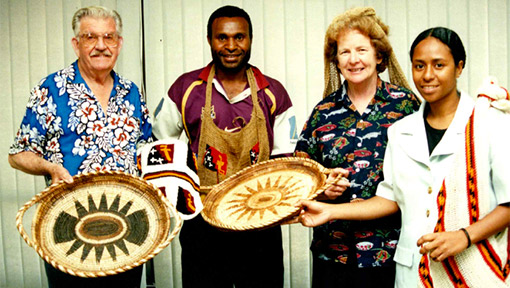 Image: “My husband Ken and I at a presentation with work colleagues, Port Moresby 2002. Traditional woven trays and bilums (string bags). I am wearing mine on the back of my head like the women do.”